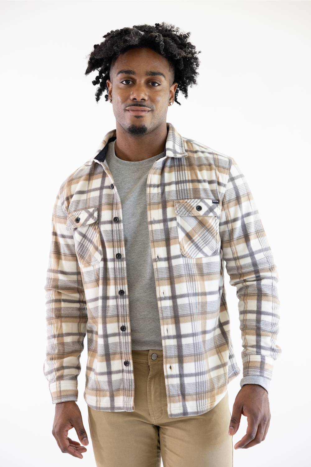A man wearing a beige, brown, and gray plaid button-up flannel shirt over a gray t-shirt and khaki pants, standing against a plain white background.