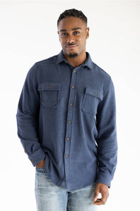 A man wearing a blue long-sleeve button-up shirt with two chest pockets and light-wash jeans, standing against a plain white studio background.