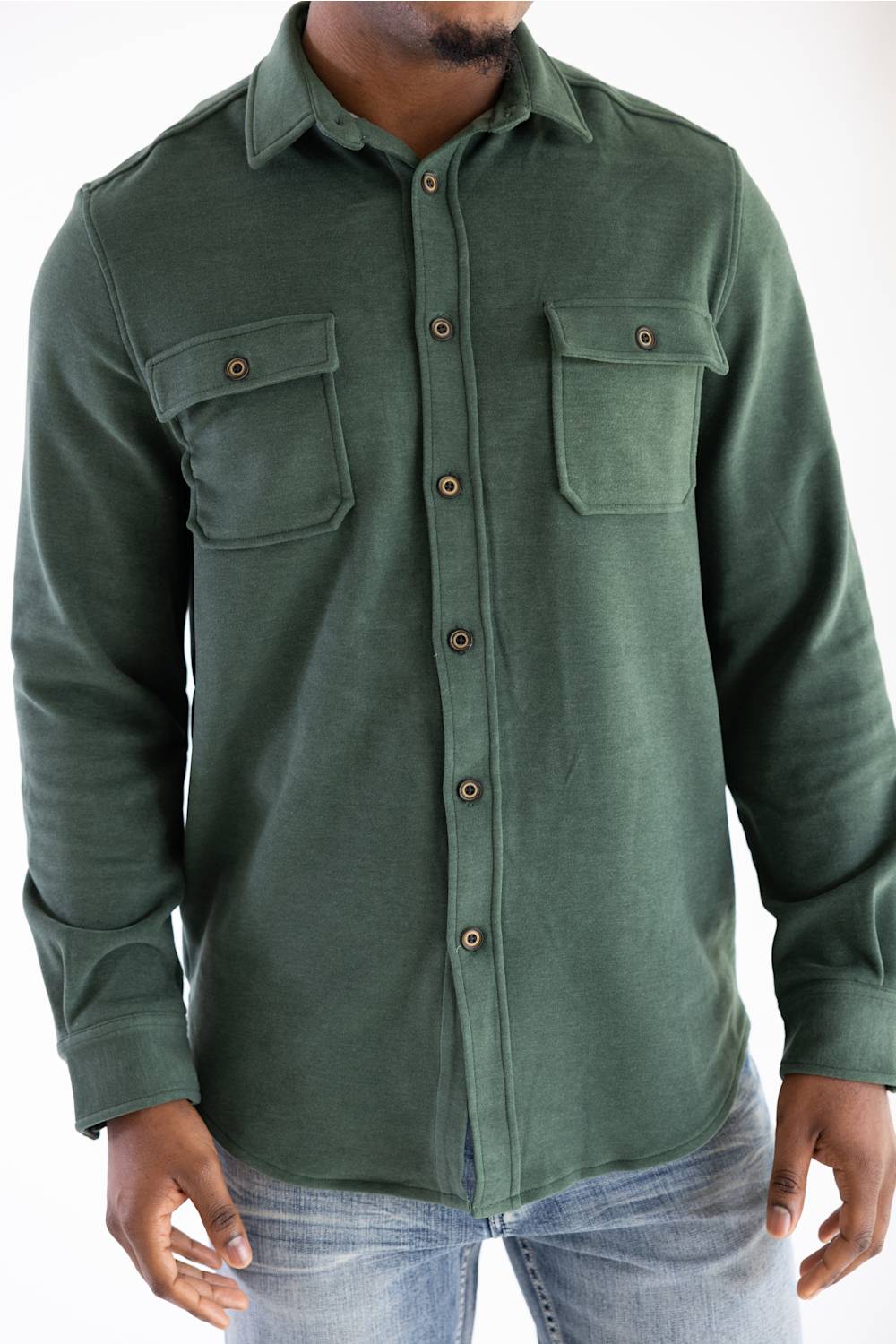 A man wearing a dark green long-sleeve button-up shirt with two chest pockets and brown buttons, paired with blue jeans, against a plain white background.