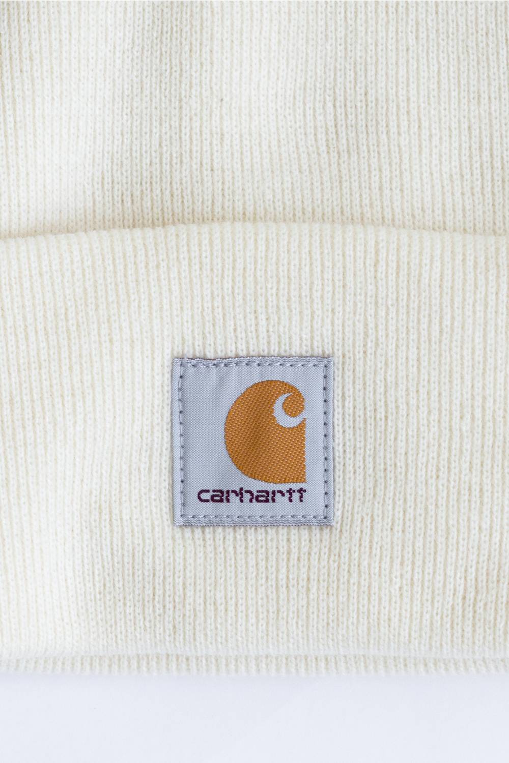 Close-up of a cream-colored ribbed knit beanie with a square Carhartt brand patch on the cuff.