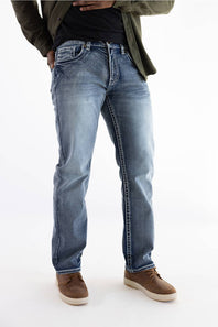 A person wearing light blue straight-leg denim jeans with a faded wash, styled with a dark olive green jacket, black shirt, and brown suede shoes against a white studio background.