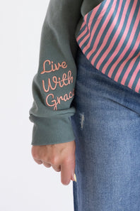 Close-up of a person wearing a sage green grey   long-sleeve top with 'Live With Grace' embroidered in pink on the sleeve, paired with a pink and navy striped shirt and blue jeans.