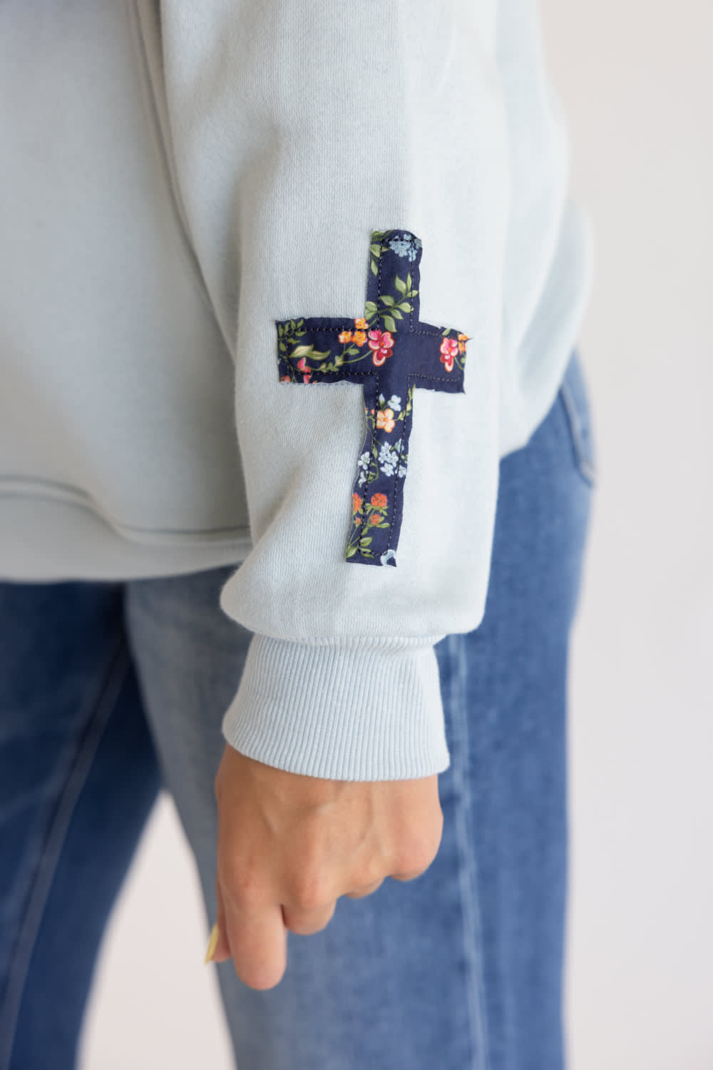 A close-up of a person wearing a light gray sweatshirt with a dark navy floral cross patch on the upper sleeve, paired with blue jeans.