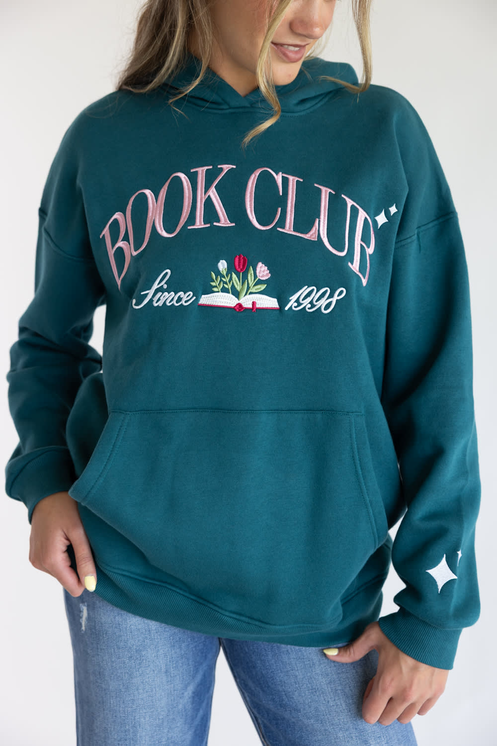 A woman wearing a teal green pullover hoodie with a graphic on the chest that reads 'BOOK CLUB Since 1998' with a book and flowers, paired with blue jeans against a plain white background.