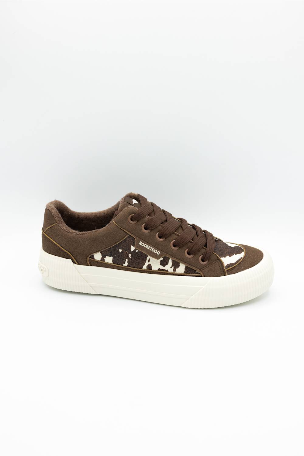 A low-top brown sneaker with a brown and white cow-print pattern on the side panel, brown laces, and a thick white platform sole, shown against a plain white background.