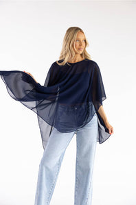 A woman modeling a sheer, flowy navy blue top with wide draped sleeves, paired with light-wash jeans, against a plain white studio background.