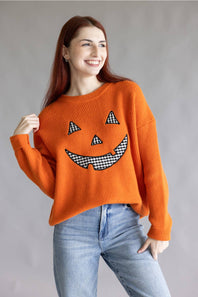 A woman wearing a bright orange ribbed knit sweater with a black-and-white gingham jack-o'-lantern face graphic, paired with light blue jeans, against a neutral gray studio background.