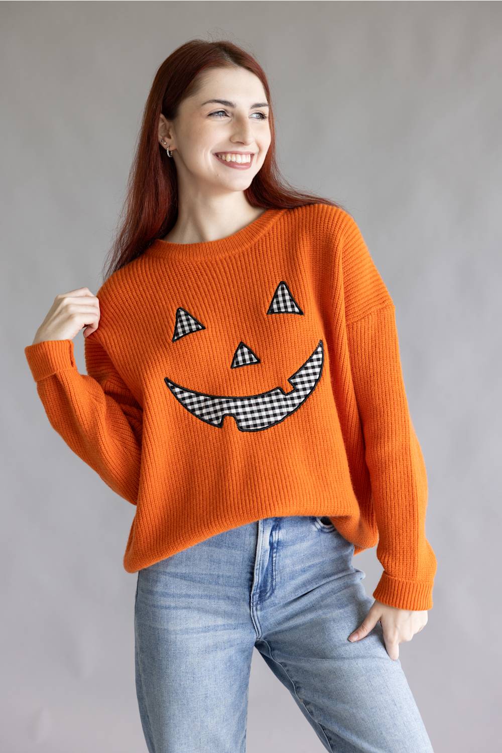 A woman wearing a bright orange ribbed knit sweater with a black-and-white gingham jack-o'-lantern face graphic, paired with light blue jeans, against a neutral gray studio background.