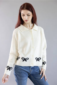 A woman wearing a cream-colored knit cardigan with black bow details on the front and cuffs, paired with blue jeans, against a solid gray studio background.