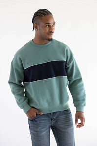 A man wearing a sage green crewneck sweatshirt with a thick navy blue horizontal stripe across the chest, paired with blue jeans, photographed against a plain white studio background.