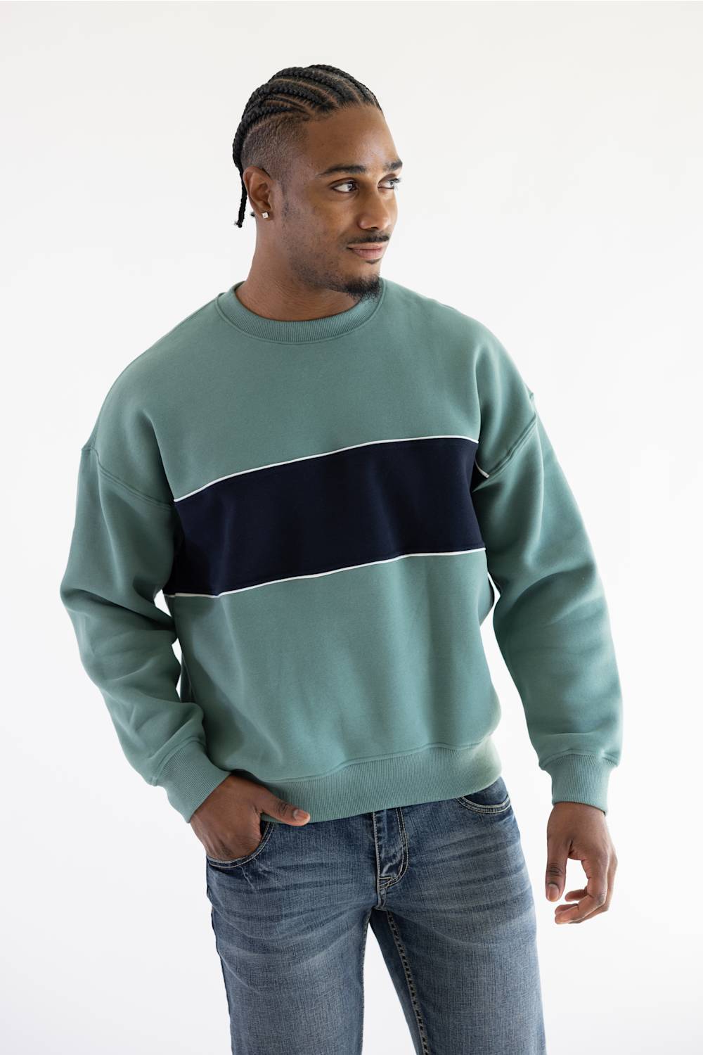 A man wearing a sage green crewneck sweatshirt with a thick navy blue horizontal stripe across the chest, paired with blue jeans, photographed against a plain white studio background.