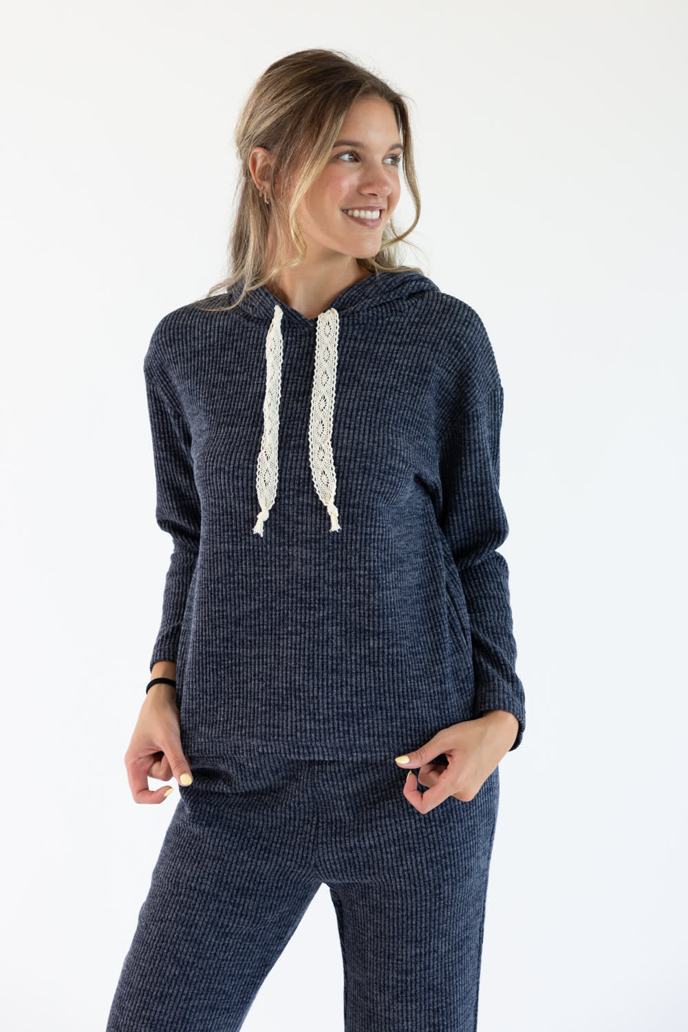 A woman modeling a dark navy blue ribbed knit loungewear set, featuring a hooded sweatshirt with cream-colored textured drawstrings and matching knit pants, against a plain white studio background.