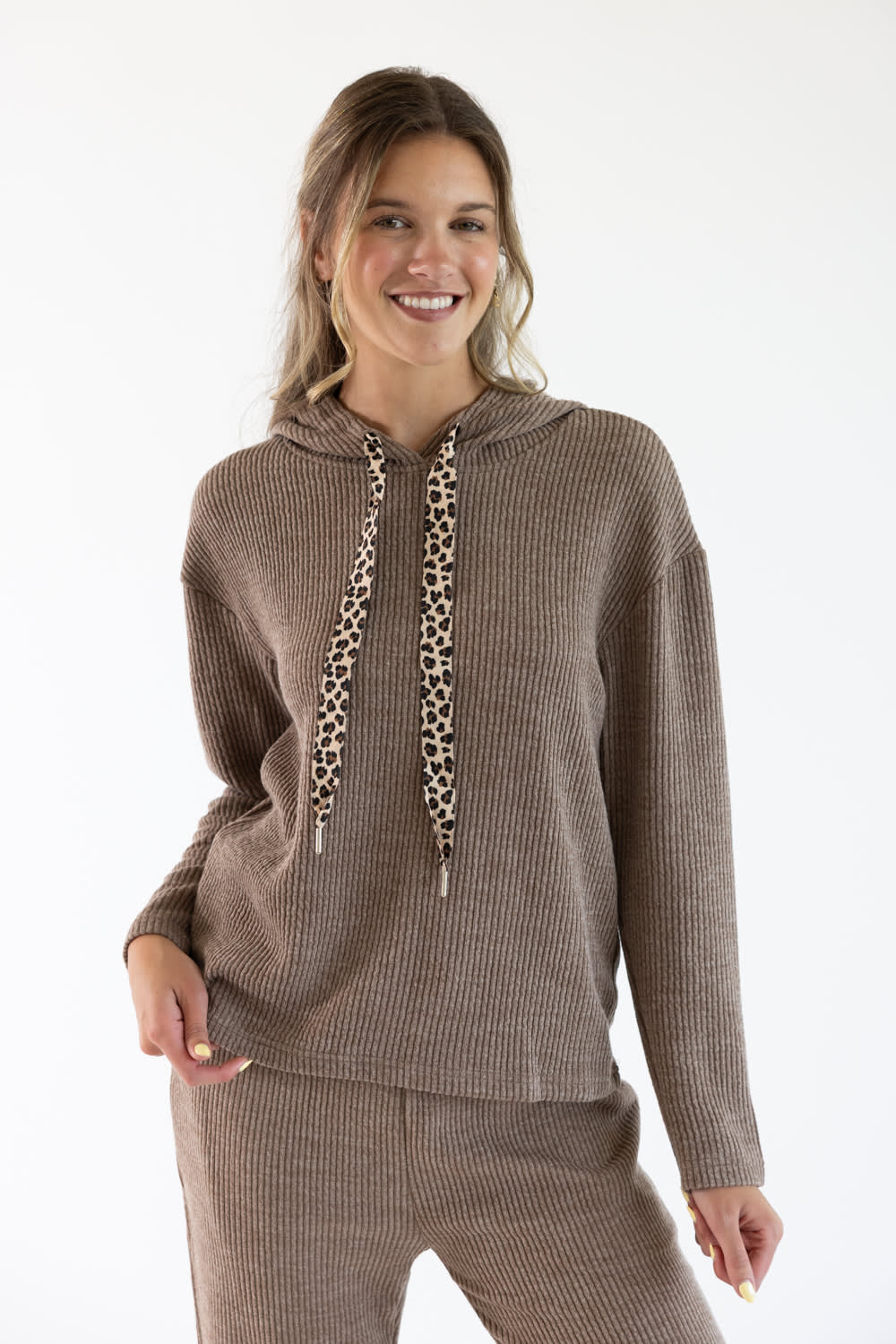 A woman modeling a ribbed knit lounge set in sand brown, featuring a hooded sweatshirt with leopard-print drawstrings and matching pants, against a plain white studio background.