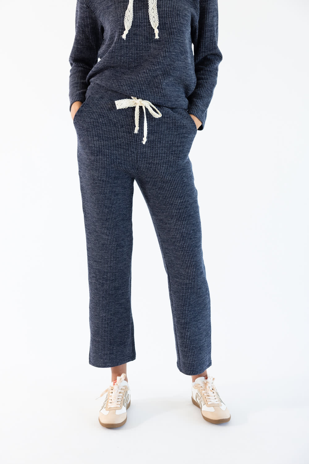 A person wearing a matching navy corduroy lounge set with a drawstring hoodie and cropped pants, paired with white sneakers, photographed against a clean white studio background.