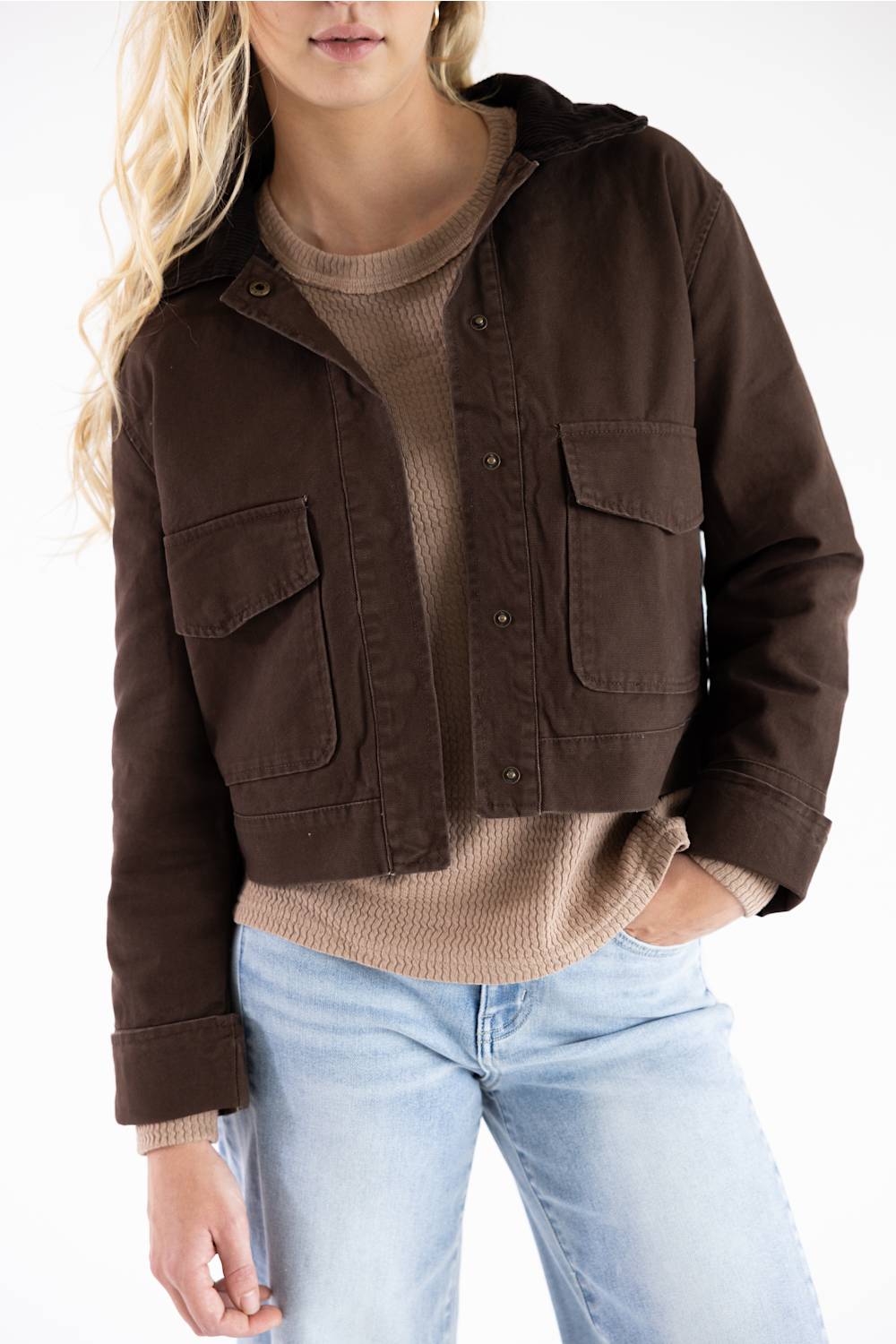A woman wearing a dark brown cropped utility jacket over a beige ribbed sweater and light blue jeans, shown from the waist up against a plain white background.