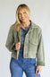 A smiling blonde woman models a sage green corduroy jacket over a blue floral shirt and blue jeans, standing against a light gray studio background.