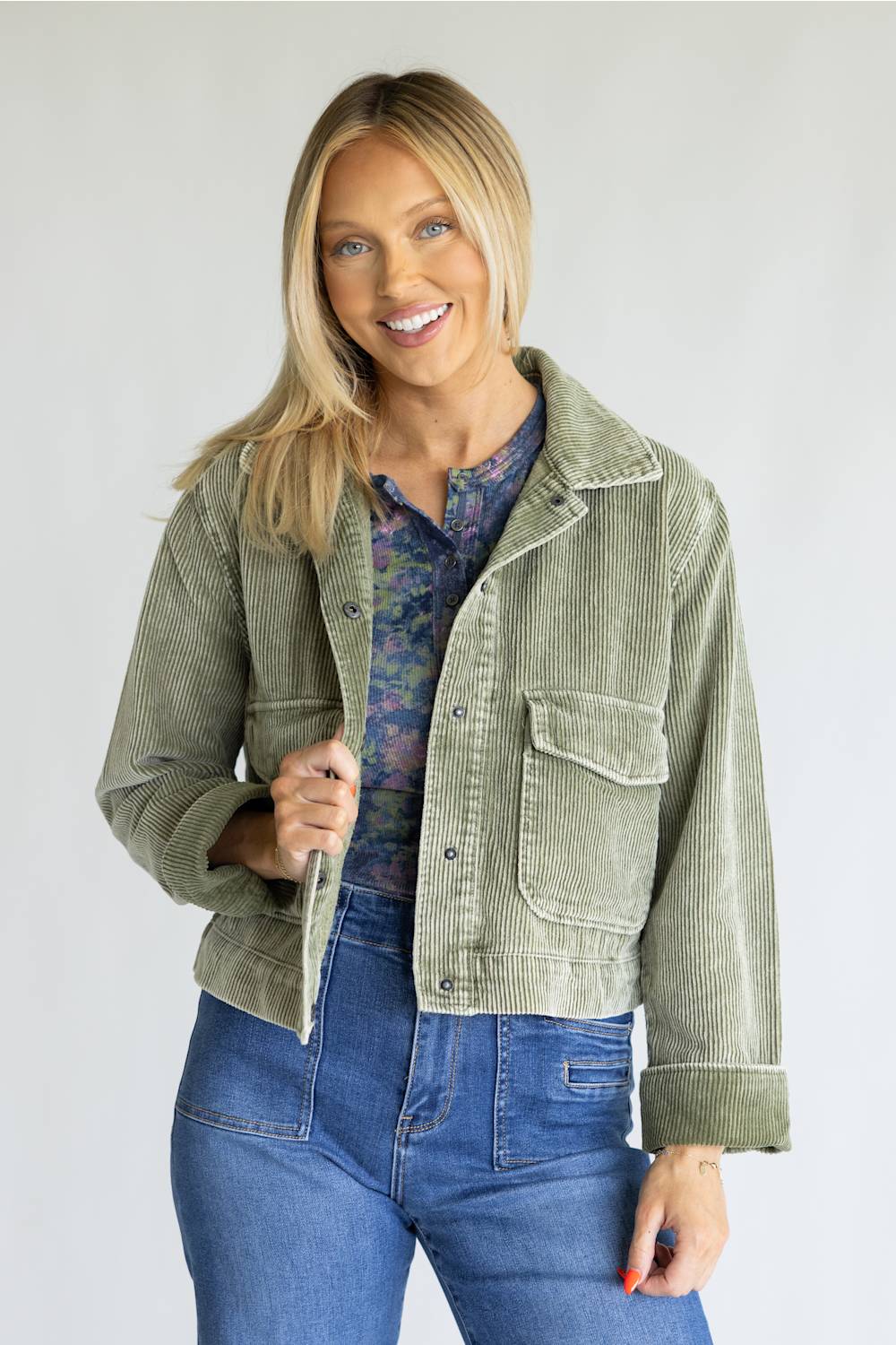 A smiling blonde woman models a sage green corduroy jacket over a blue floral shirt and blue jeans, standing against a light gray studio background.