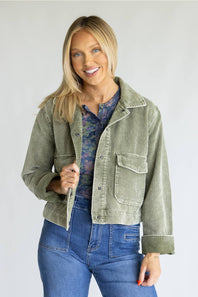 A smiling blonde woman models a sage green corduroy jacket over a blue floral shirt and blue jeans, standing against a light gray studio background.