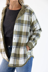 A woman wearing an oversized plaid flannel button-up shirt in olive green, white, and blue, layered over a dark gray knit sweater and blue jeans.