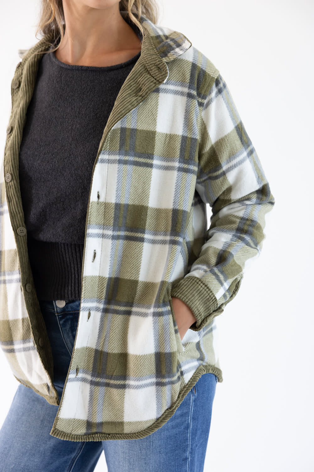 A woman wearing an oversized plaid flannel button-up shirt in olive green, white, and blue, layered over a dark gray knit sweater and blue jeans.