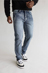 A person wearing light-wash blue denim jeans with a faded wash, styled with a black long-sleeve top and black-and-white sneakers, standing against a plain light gray studio background.