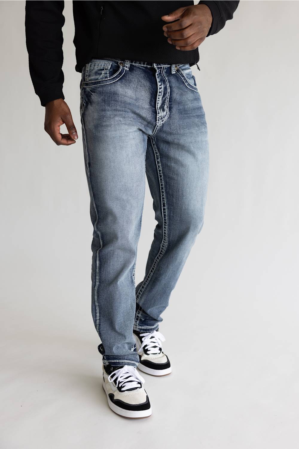 A person wearing light-wash blue denim jeans with a faded wash, styled with a black long-sleeve top and black-and-white sneakers, standing against a plain light gray studio background.