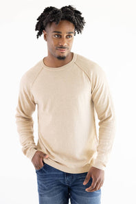 A man wearing a light beige knit crewneck sweater with raglan sleeves and blue jeans, standing against a plain white background.