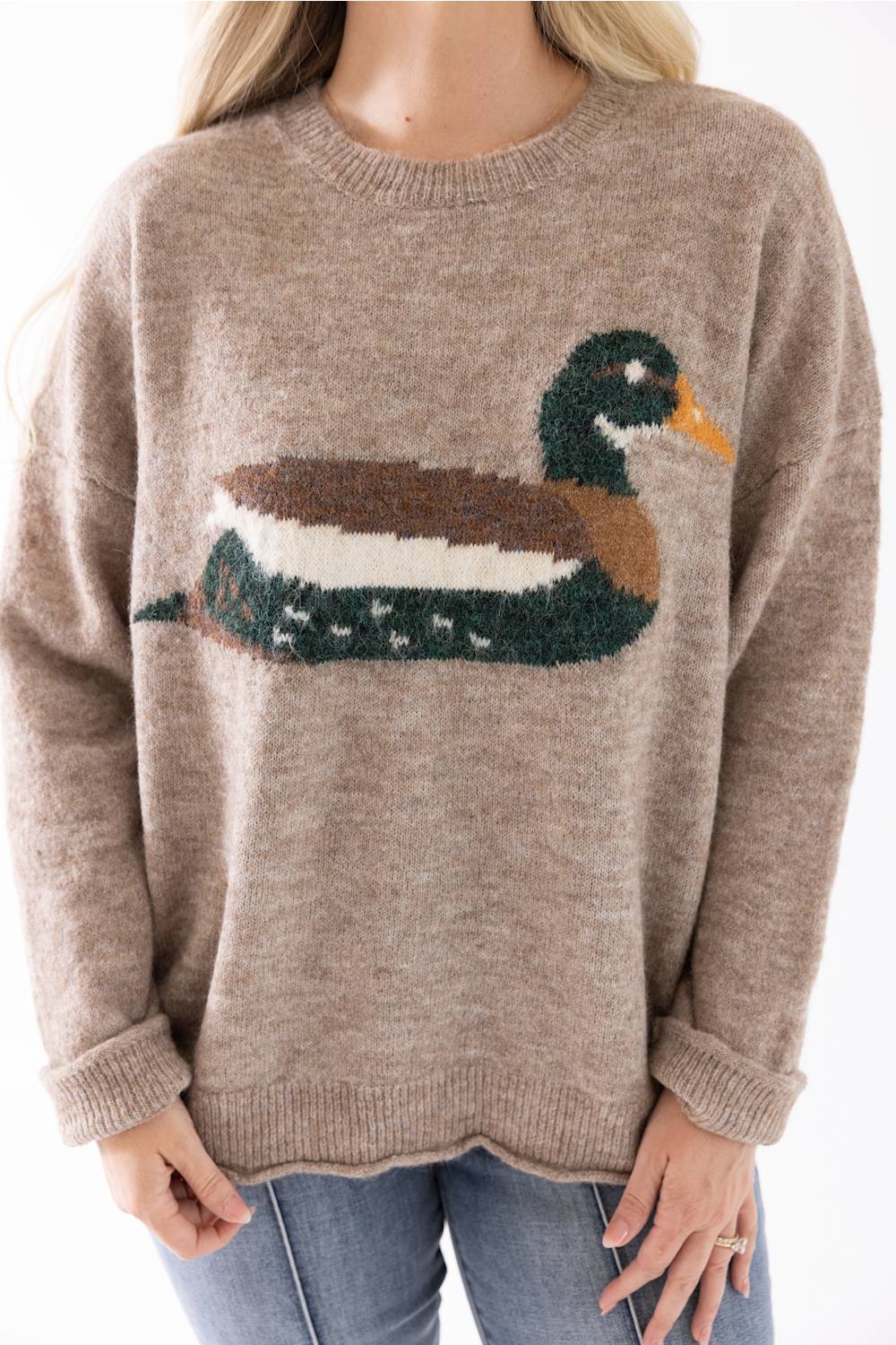 A woman wearing a taupe knit sweater with a large duck motif across the chest, paired with blue jeans, against a plain white background.