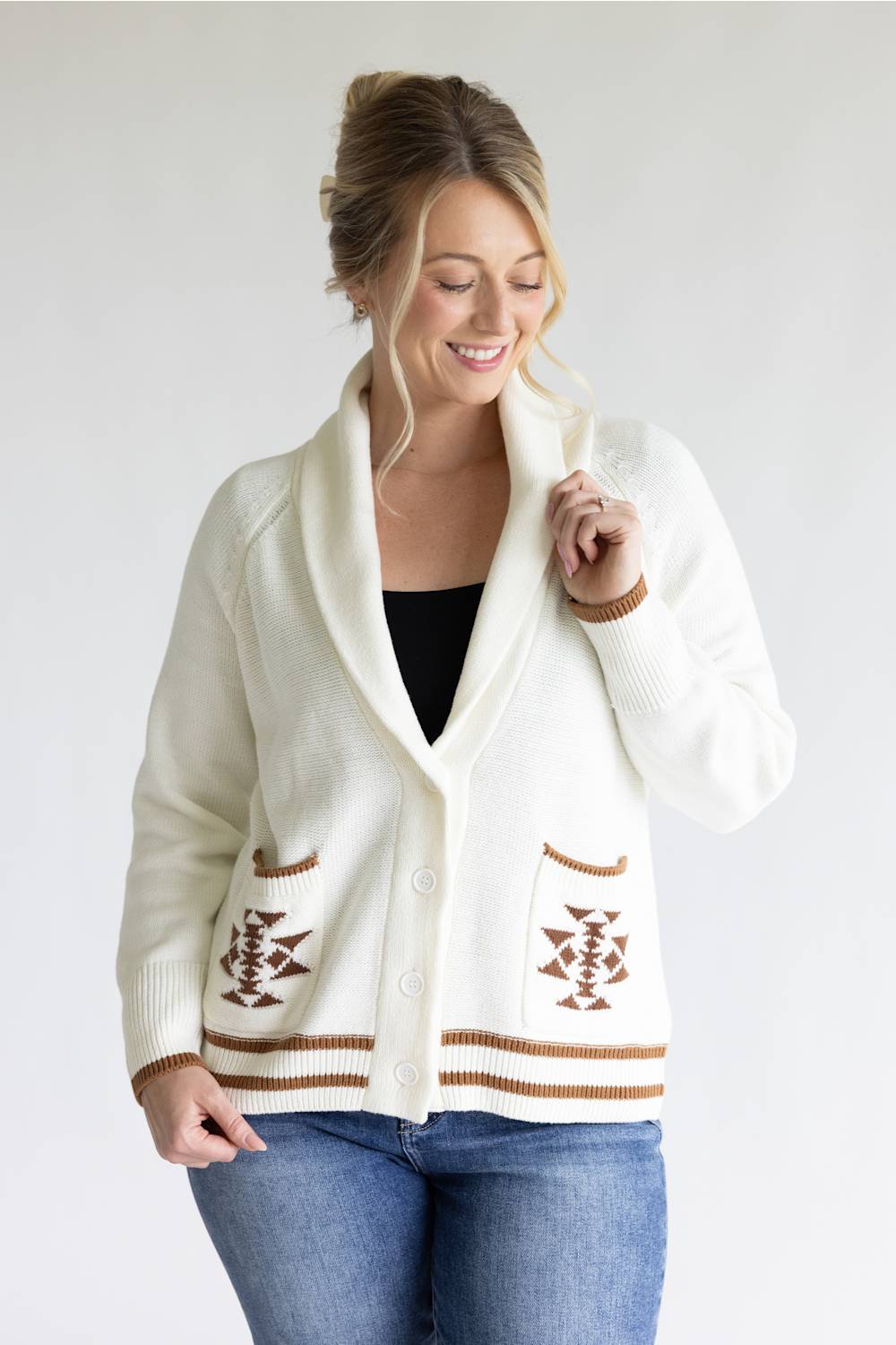 A woman wearing a cream-colored knit cardigan with brown geometric patterns and striped trim, styled over a black top and blue jeans, against a plain light gray background.