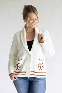 A woman wearing a cream-colored knit cardigan with brown geometric patterns and striped trim, styled over a black top and blue jeans, against a plain light gray background.