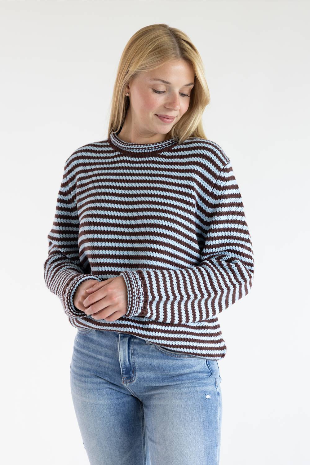 A woman wearing a long-sleeve knit sweater with horizontal brown, black, and white stripes, paired with light blue jeans, photographed against a plain white studio background.