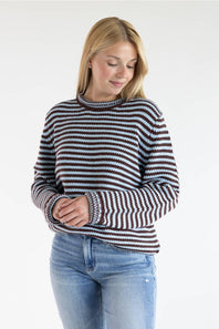 A woman wearing a long-sleeve knit sweater with horizontal brown, black, and white stripes, paired with light blue jeans, photographed against a plain white studio background.