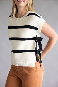A woman wearing a cream ribbed knit sleeveless sweater vest with black horizontal stripes and a black side bow, paired with brown pants, against a neutral gray background.