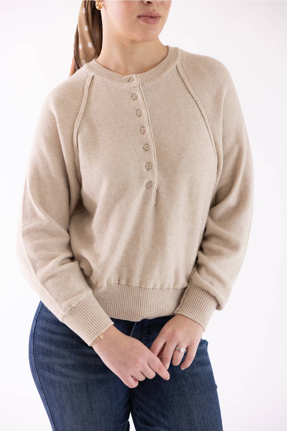 A woman wearing a beige knit button-front sweater with raglan sleeves and ribbed collar, cuffs, and hem, paired with dark blue jeans against a plain white background.