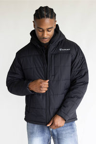 A man wearing a black Ariat quilted hooded jacket with a white logo on the chest, paired with blue jeans, photographed against a plain light background.