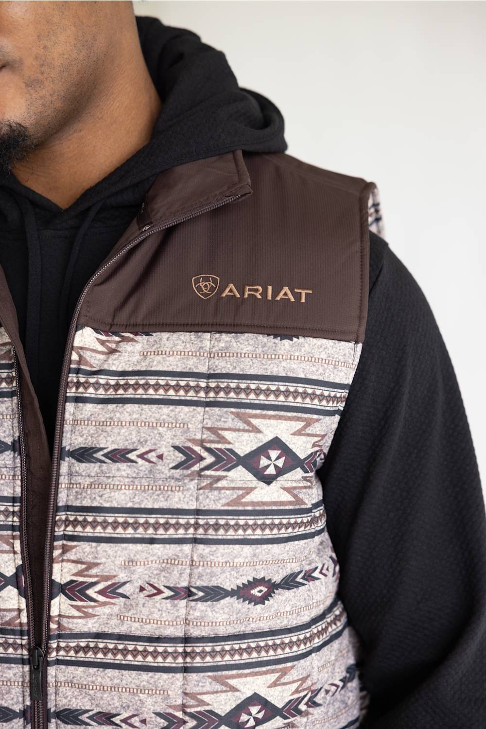 A close-up of a person wearing an Ariat quilted vest with a brown and geometric patterned design, layered over a black hoodie.