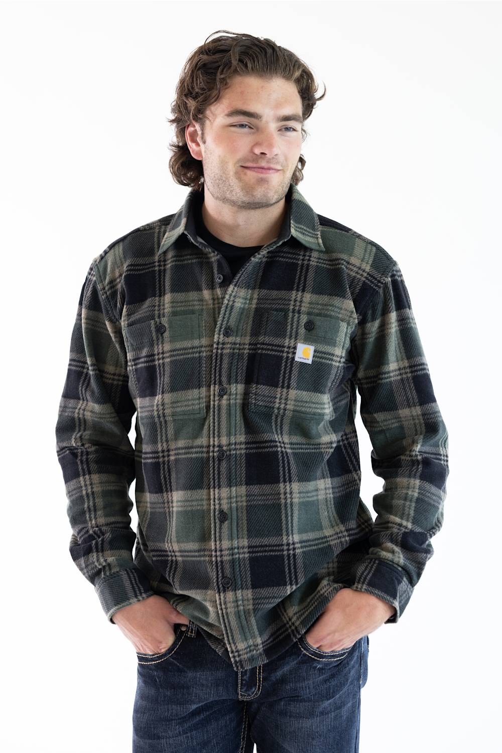 A man wearing a Carhartt long-sleeve plaid button-up shirt in dark green, black, and gray, paired with blue jeans, against a plain white background.