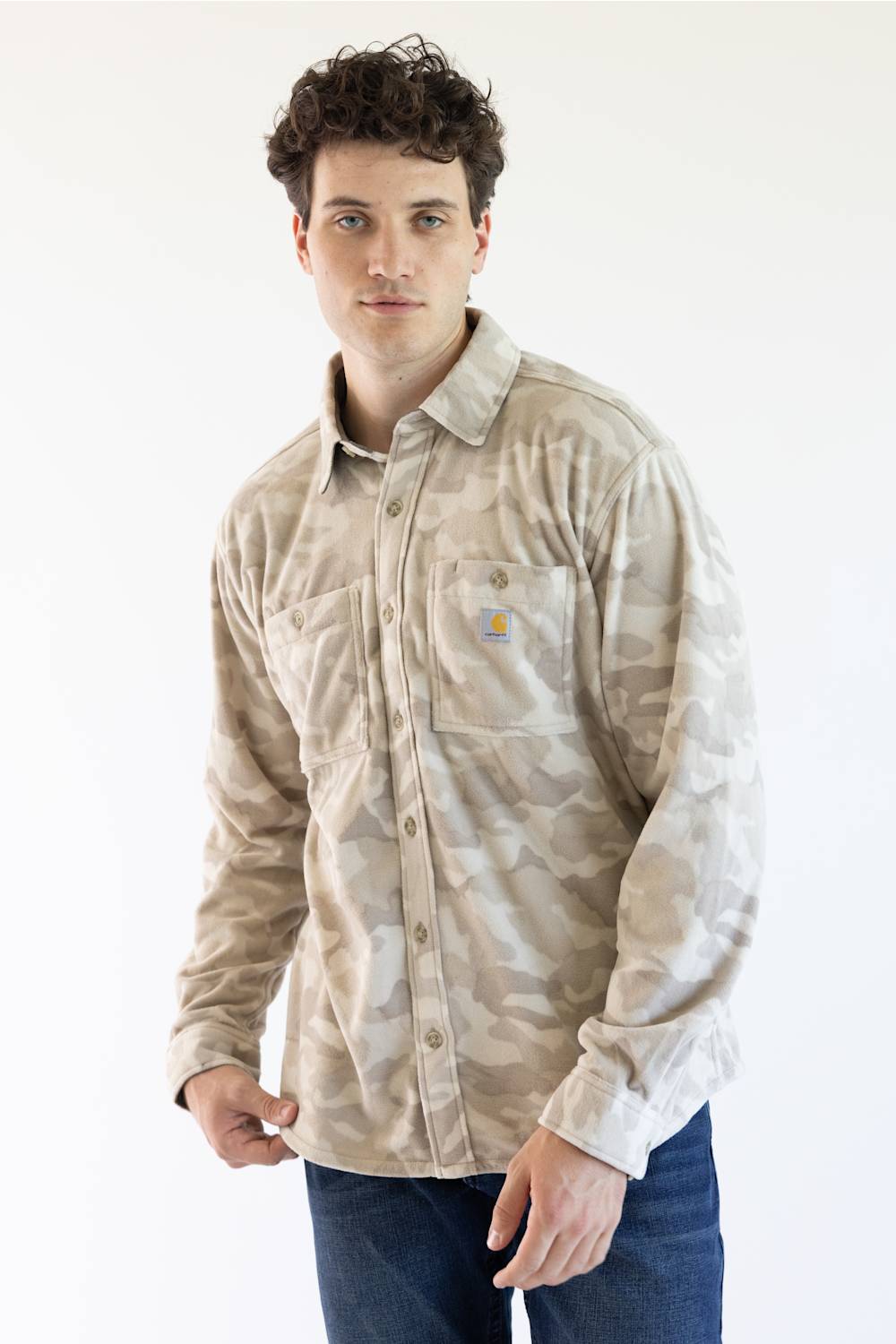 A man wearing a beige and light brown camouflage long-sleeve button-up shirt with a chest pocket, paired with blue jeans, standing against a plain white background.