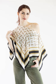 A woman modeling a white scarf with a repeating gold monogram pattern and striped borders in black, navy, and gold, styled over her shoulder.