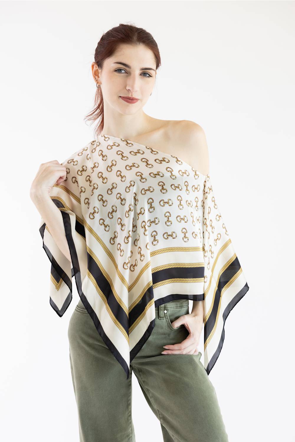 A woman modeling a white scarf with a repeating gold monogram pattern and striped borders in black, navy, and gold, styled over her shoulder.