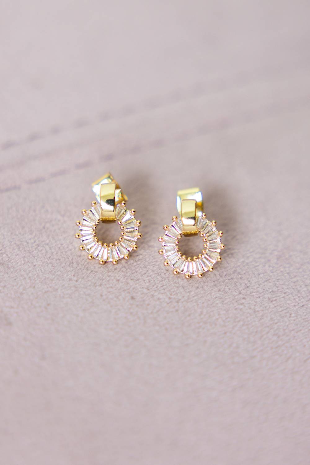 A pair of gold hoop earrings with clear faceted baguette crystals arranged in a circular halo design, resting on a textured light beige surface.