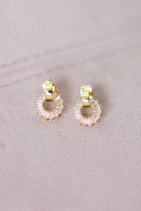 A pair of gold hoop earrings with clear faceted baguette crystals arranged in a circular halo design, resting on a textured light beige surface.