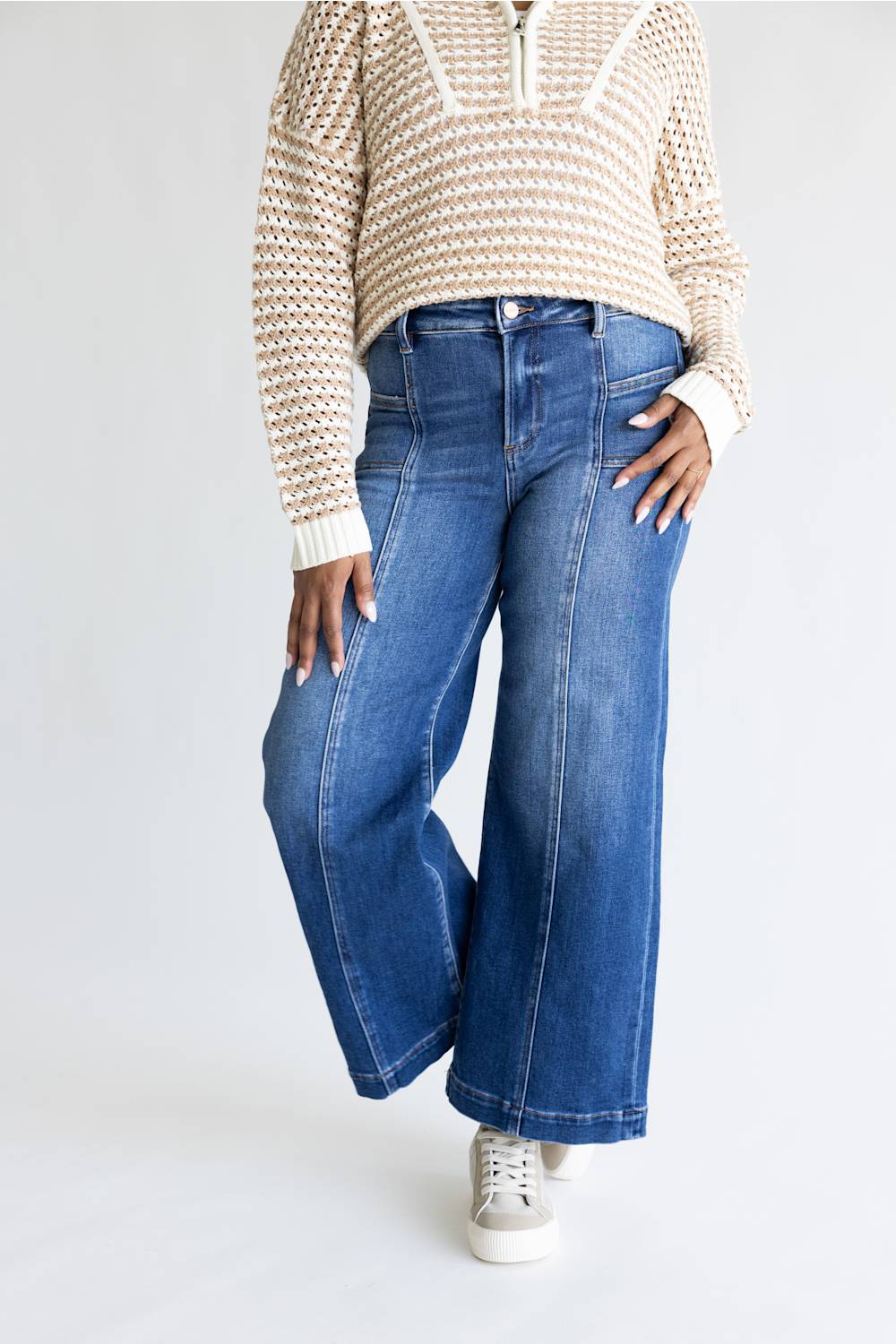 A model wearing a pair of wide-leg blue denim jeans with a textured knit sweater and white sneakers, shown from the chest down against a plain white studio background.