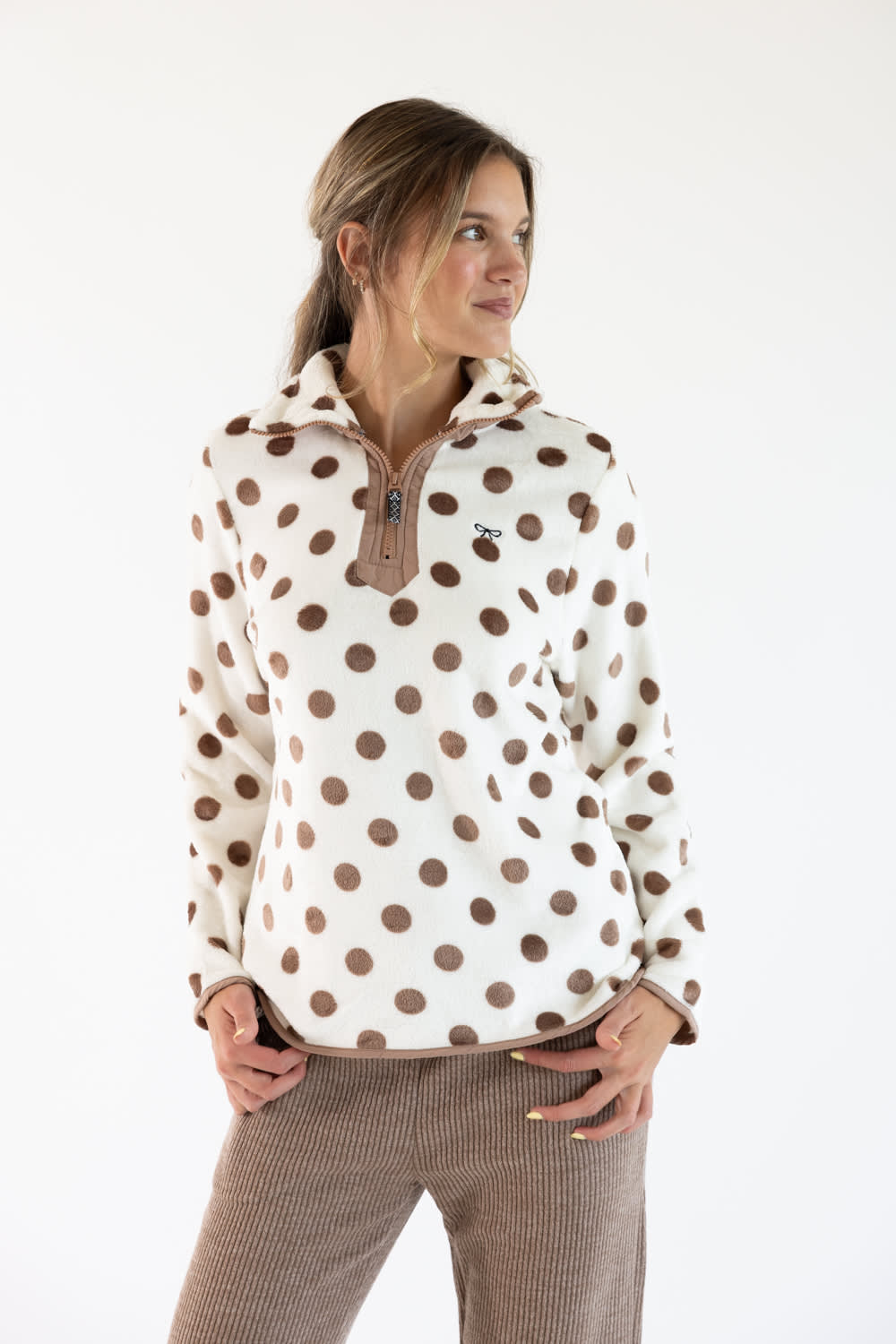 A woman modeling a cream fleece pullover with brown polka dots and a brown zipper collar, paired with brown corduroy pants, against a plain white background.
