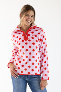 A woman modeling a pink hooded quarter-zip fleece pullover with red polka dots and red trim, paired with blue jeans, against a plain white studio background.