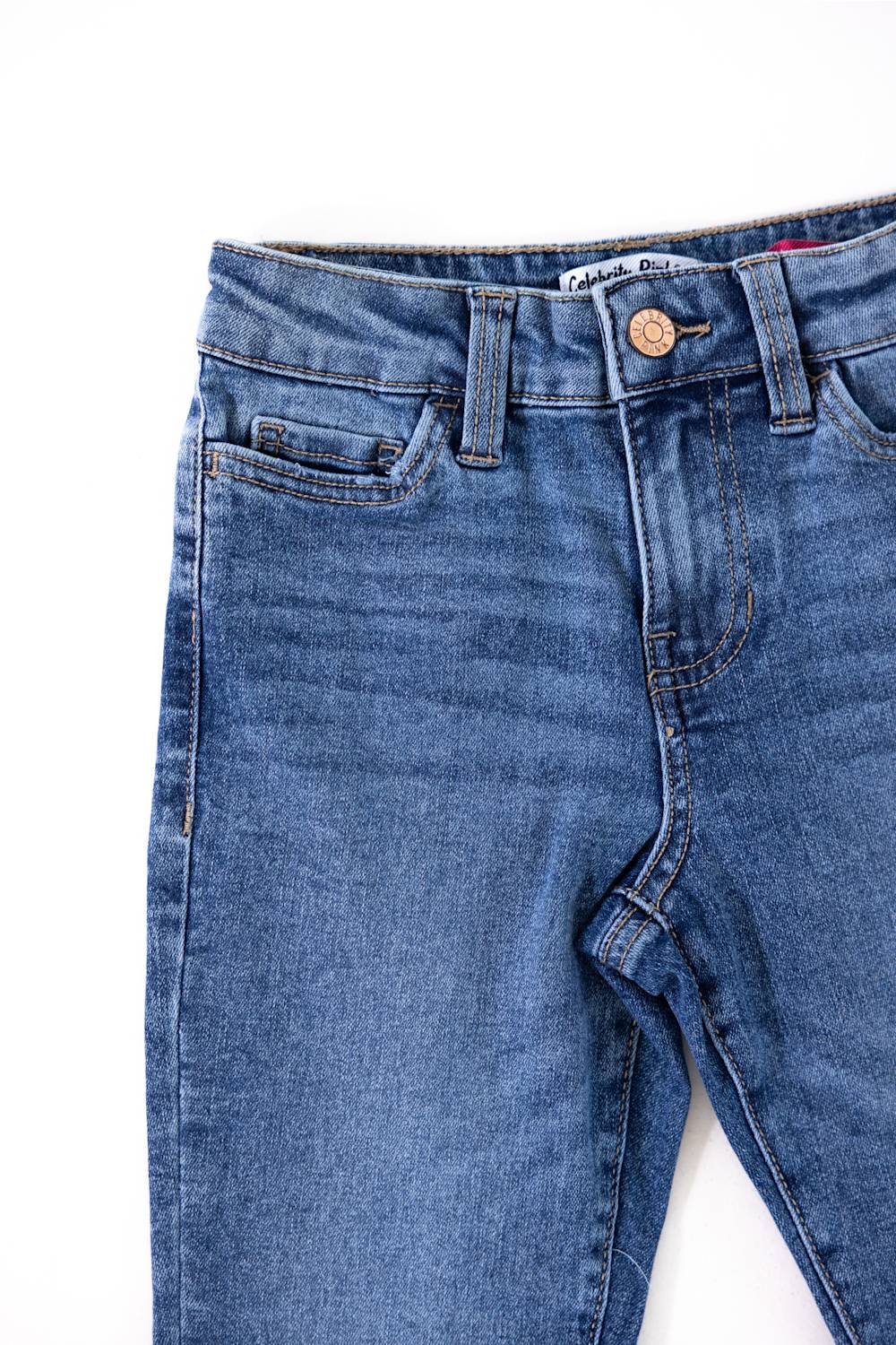Close-up view of the waistband and front pockets of Celebrity Pink girls medium wash denim jeans, featuring a copper button, zip fly, belt loops, and subtle whiskering.