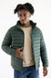 A man wearing a green Patagonia quilted puffer jacket over a beige sweater and blue jeans, standing against a plain white background.