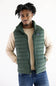 A man wearing a Patagonia green quilted puffer vest over a beige long-sleeve shirt and blue jeans, standing against a plain white background.