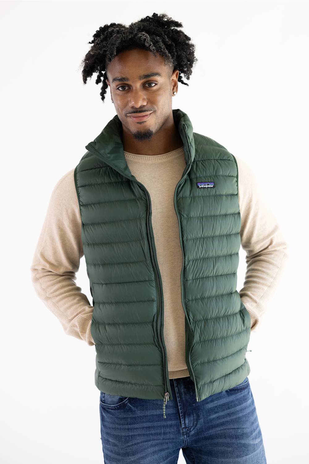 A man wearing a Patagonia green quilted puffer vest over a beige long-sleeve shirt and blue jeans, standing against a plain white background.
