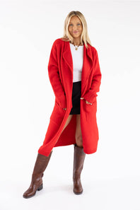 A woman modeling a bright red open-front knit cardigan/coat over a white top and black skirt, paired with brown knee-high boots, against a plain white studio background.